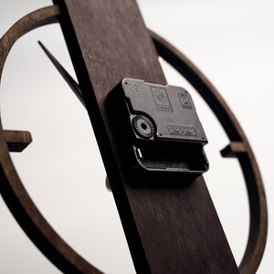 May include: Close-up of a wooden clock with a dark brown finish. The clock features a circular design with a rectangular base. The clock mechanism is visible, with the word "QUARTZ" printed on it. The clock hands are black.