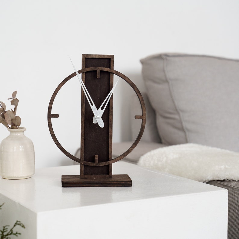 May include: A modern wooden desk clock with a dark brown finish. The clock features a circular frame with minimalist hour markers and white clock hands. The clock is approximately 10 inches tall and sits on a rectangular base.