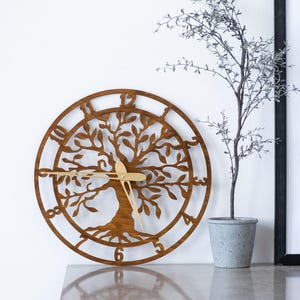 May include: A circular wooden wall clock featuring a laser-cut tree of life design with intricate branches and roots. The clock face has large cutout numerals from one to twelve along the outer ring and thin, gold-colored metal clock hands.
