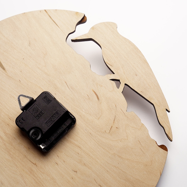 May include: A wooden wall clock with a cutout design of a bird perched on a branch. The clock face is light brown, and the clock mechanism is black. The clock has a unique, natural aesthetic.