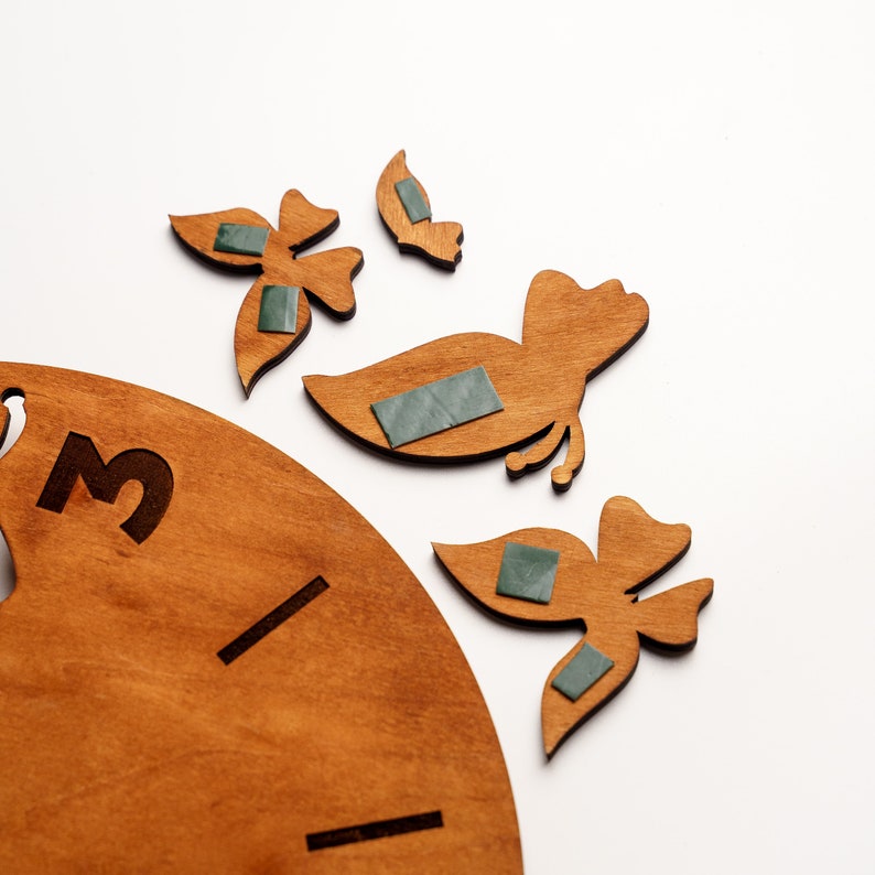 May include: Wooden clock face with the number 3 and several wooden bird and butterfly cutouts. The cutouts have adhesive squares on the back. The clock face and cutouts are a light brown color.