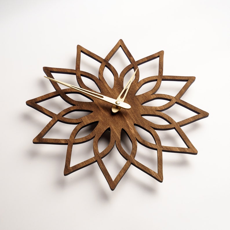 K&ouml;nnte beinhalten: Eine Wanduhr aus Holz mit floralem Design. Das Zifferblatt der Uhr ist dunkelbraun, mit einem bl&uuml;tenartigen Muster aus dem Holz geschnitten. Die Zeiger der Uhr sind goldfarben.