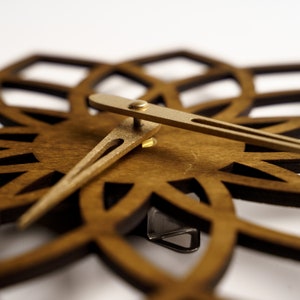 Puede incluir: Primer plano de un reloj de pared de madera con dise&ntilde;o floral. El reloj presenta una esfera de madera marr&oacute;n oscuro con patrones recortados y manecillas doradas. Las manecillas del reloj indican la hora.
