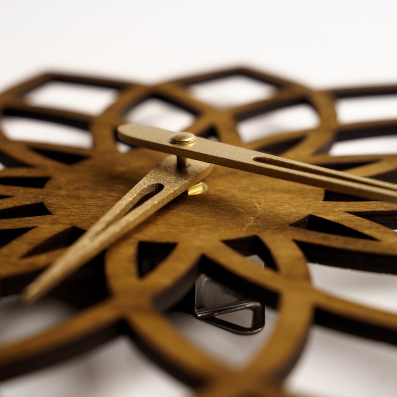 Puede incluir: Primer plano de un reloj de pared de madera con dise&ntilde;o floral. El reloj presenta una esfera de madera marr&oacute;n oscuro con patrones recortados y manecillas doradas. Las manecillas del reloj indican la hora.
