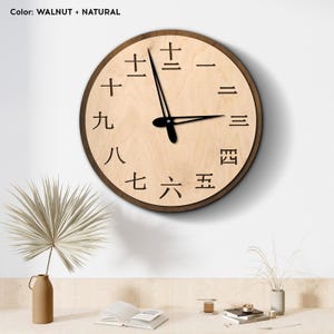 Pode incluir: Um relógio de parede redondo de madeira com moldura cor de nogueira e mostrador de madeira natural. O relógio apresenta ponteiros pretos e algarismos japoneses pretos. O texto "Color: WALNUT + NATURAL" está no topo.