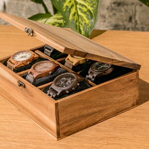 Pode incluir: Uma caixa de rel&oacute;gio de madeira com tampa articulada, exibindo seis rel&oacute;gios com v&aacute;rios designs de pulseira e mostrador. A caixa &eacute; feita de madeira marrom clara e tem um interior escuro.