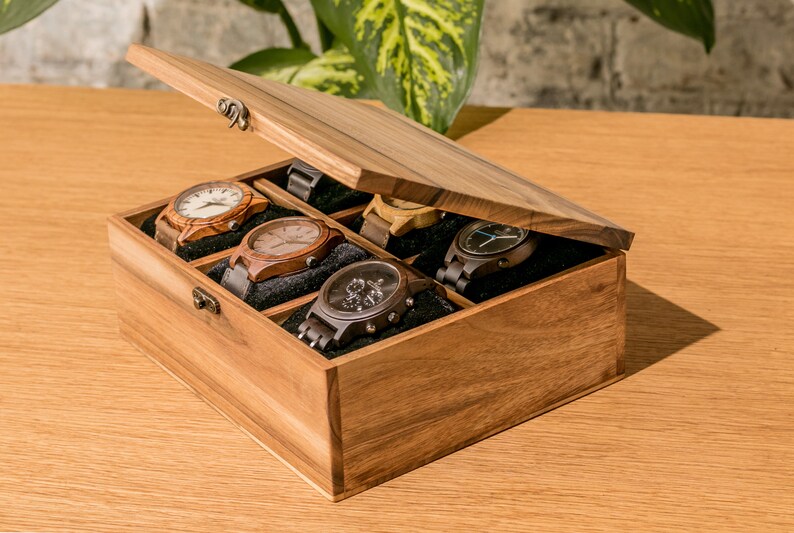Pode incluir: Uma caixa de rel&oacute;gio de madeira com tampa articulada, exibindo seis rel&oacute;gios com v&aacute;rios designs de pulseira e mostrador. A caixa &eacute; feita de madeira marrom clara e tem um interior escuro.