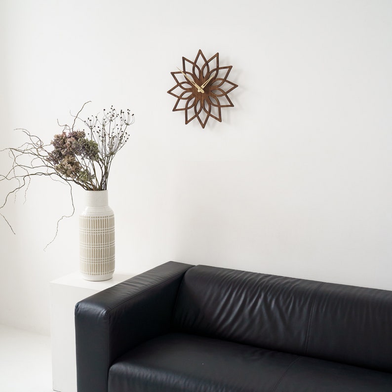 K&ouml;nnte beinhalten: Eine braune Wanduhr aus Holz in floralem Design mit goldenen Zeigern, an einer wei&szlig;en Wand befestigt. Eine Vase mit getrockneten Blumen steht auf einer wei&szlig;en Oberfl&auml;che, und ein schwarzes Ledersofa befindet sich im Vordergrund. Das Design der Uhr &auml;hnelt einer Blume.