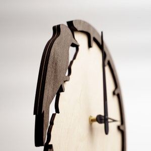 May include: Wooden wall clock with a bird silhouette cut-out design. The clock features a light-colored face with dark brown edging and a bird shape on the left side. The clock hands are black, and the central post is gold.