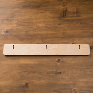 May include: A light-colored wooden wall-mounted rack with three evenly spaced holes for hanging. The rack is rectangular with rounded corners and is set against a brown wooden background.