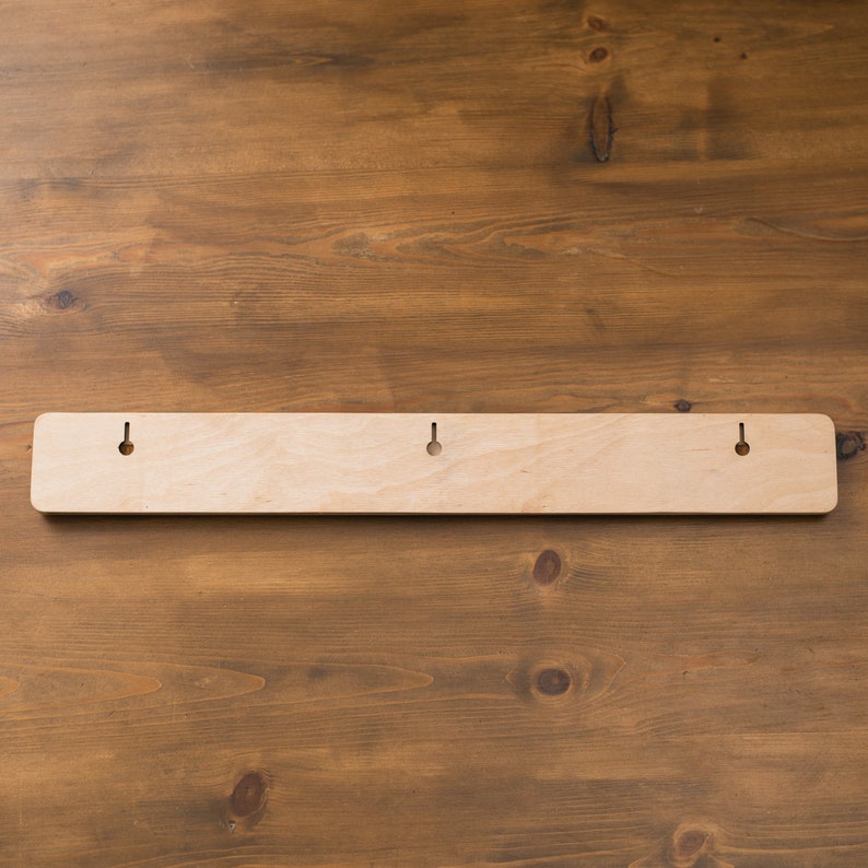 May include: A light-colored wooden wall-mounted rack with three evenly spaced holes for hanging. The rack is rectangular with rounded corners and is set against a brown wooden background.