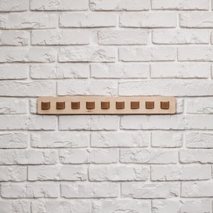 May include: A wooden wall-mounted coat rack with ten brown square pegs. The rack is made of light-colored wood and is mounted on a white brick wall. The pegs are evenly spaced along the rack.