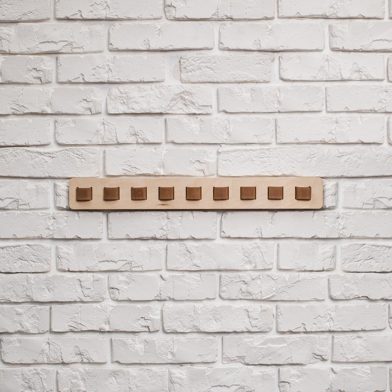 May include: A wooden wall-mounted coat rack with ten brown square pegs. The rack is made of light-colored wood and is mounted on a white brick wall. The pegs are evenly spaced along the rack.