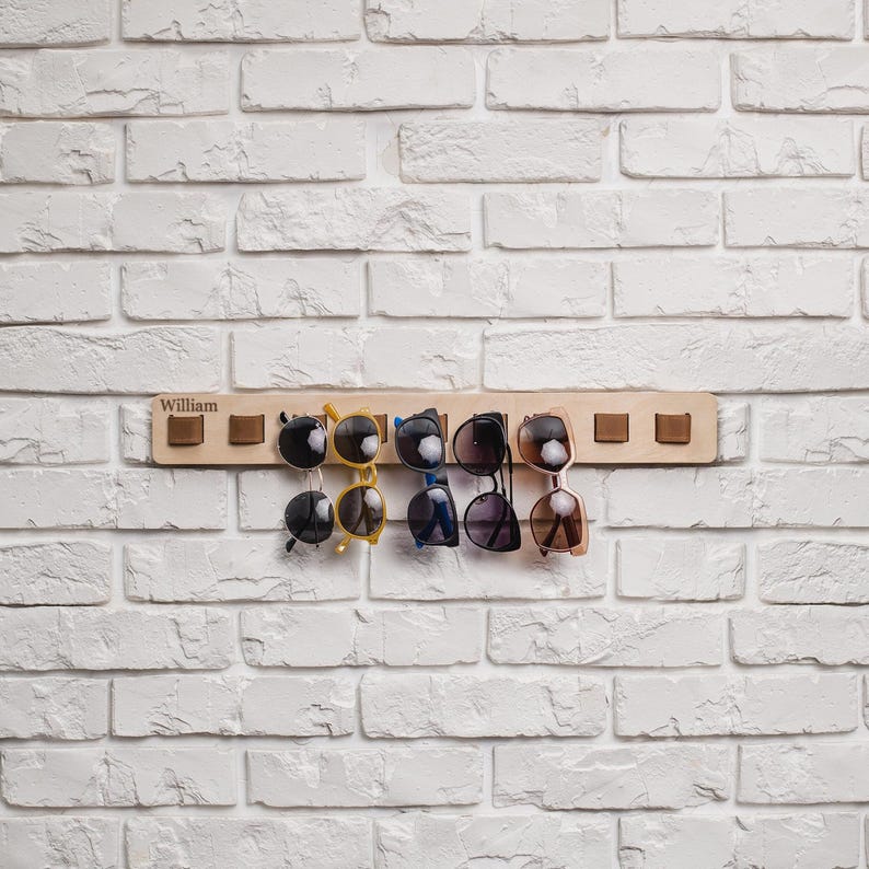 A wooden wall-mounted sunglasses holder labeled “William” holds six pairs of sunglasses by leather loops. The holder is attached to a white-painted brick wall and has eight evenly spaced loops, with two empty on each end. The sunglasses vary in shape and color, including black, yellow, blue, and brown frames.