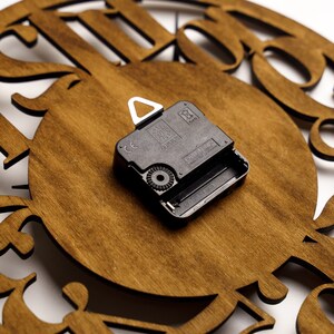 Puede incluir: Primer plano de una esfera de reloj de madera con un movimiento de reloj de cuarzo negro. La esfera del reloj es de color marr&oacute;n c&aacute;lido con dise&ntilde;os decorativos recortados. El movimiento del reloj es negro y tiene un colgador de metal blanco.