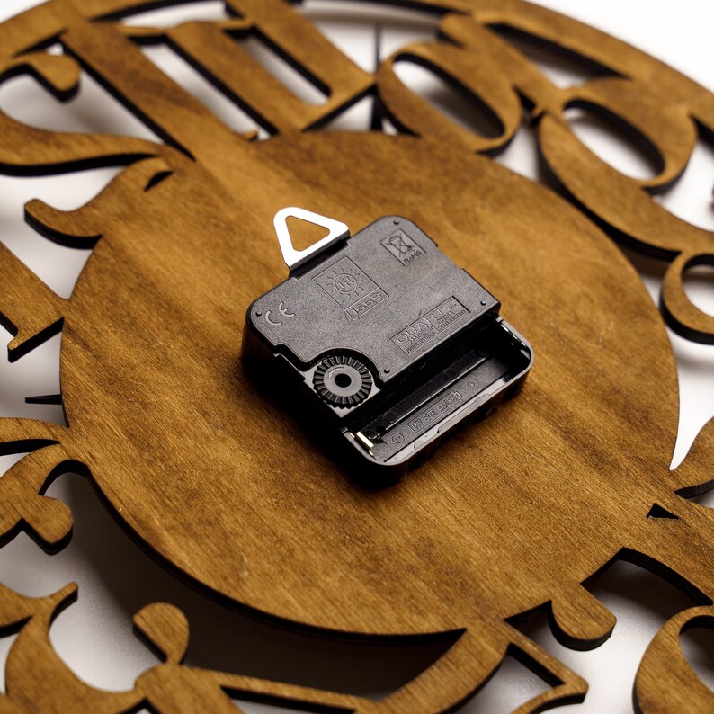 Puede incluir: Primer plano de una esfera de reloj de madera con un movimiento de reloj de cuarzo negro. La esfera del reloj es de color marr&oacute;n c&aacute;lido con dise&ntilde;os decorativos recortados. El movimiento del reloj es negro y tiene un colgador de metal blanco.