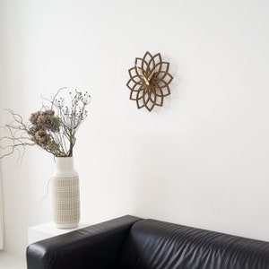 Puede incluir: Un reloj de pared de madera marr&oacute;n con dise&ntilde;o floral cuelga de una pared blanca. El reloj tiene manecillas doradas. Un jarr&oacute;n blanco con un patr&oacute;n beige contiene flores secas. Un sof&aacute; de cuero negro est&aacute; en primer plano. El di&aacute;metro del reloj es de unos 30 cm.