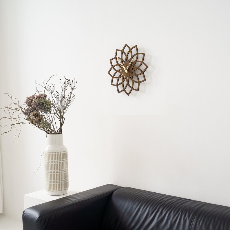 Puede incluir: Un reloj de pared de madera marr&oacute;n con dise&ntilde;o floral cuelga de una pared blanca. El reloj tiene manecillas doradas. Un jarr&oacute;n blanco con un patr&oacute;n beige contiene flores secas. Un sof&aacute; de cuero negro est&aacute; en primer plano. El di&aacute;metro del reloj es de unos 30 cm.