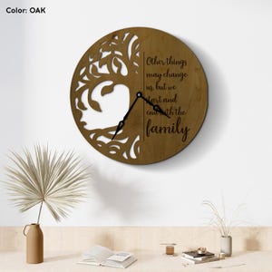 Puede incluir: Reloj de pared redondo color roble con un diseño de árbol recortado en el lado izquierdo. El reloj presenta manecillas negras y el texto "Other things may change us, but we start and end with the family."