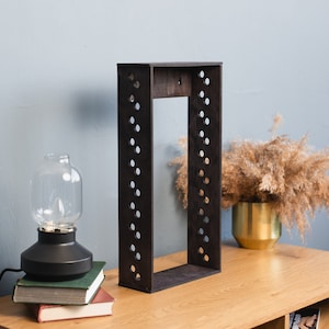 Pode incluir: Prateleira de madeira castanha escura com estrutura retangular e recortes circulares nas laterais. A prateleira está sobre uma superfície de madeira, com uma lâmpada e livros à esquerda e um vaso decorativo à direita. Aproximadamente 60 cm de altura.