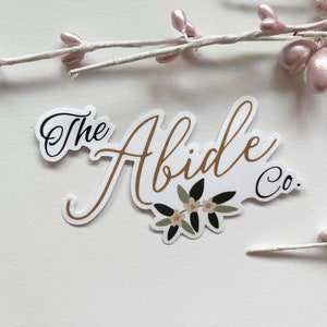Puede incluir: Pegatina blanca con letras doradas que dicen "The Abide Co." con un diseño floral en el centro. La pegatina tiene un fondo blanco y letras negras.