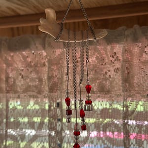 May include: A hanging ornament featuring a curved, off-white bone structure suspended by a silver-toned metal chain. Multiple silver chains dangle from the bone, each adorned with a vertical arrangement of clear and vibrant red glass beads.