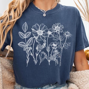 May include: A navy blue t-shirt featuring a white floral design. The graphic includes various flowers and leaves. The shirt is paired with denim shorts and a cream-colored cardigan. The word "DENIM" is visible in the bottom left corner.