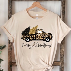 Op de afbeelding: Een beige T-shirt met een zwarte en gouden luipaardprint truck die een kerstboom vervoert. De tekst "Merry Christmas" is onder de truck gedrukt met een hartje.