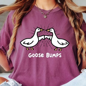 May include: A woman wearing a purple t-shirt with a white goose design and the text "Goose Bumps". She is sitting on a bed with her legs crossed.