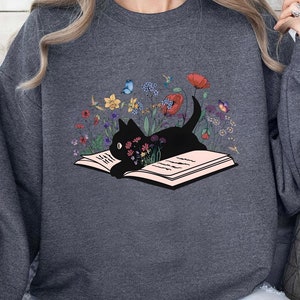 May include: A dark gray sweatshirt with a black cat reading a book surrounded by flowers and butterflies. The cat is lying on the open book, which is pink with white pages.