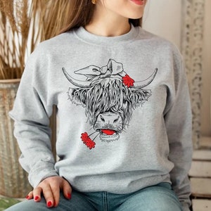 May include: A gray sweatshirt with a black and white illustration of a Highland cow wearing a red bandana and holding red flowers in its mouth.