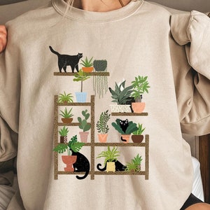 May include: A beige sweatshirt with a graphic print of black cats and potted plants on a wooden shelf. The graphic is a whimsical illustration of a cat-filled plant room.