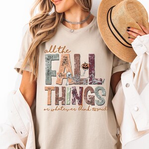 May include: A sand-colored t-shirt featuring a graphic print with the text all the FALL THINGS or whatever blink 182 said. The design includes distressed lettering in muted tones, a small skeleton figure, and a pumpkin illustration.