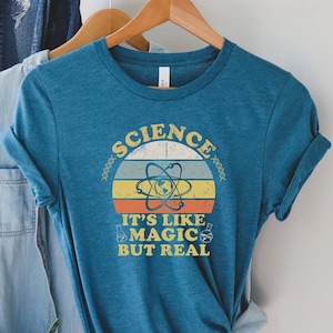 May include: A teal blue t-shirt with a vintage-style graphic that says "Science It's Like Magic But Real". The graphic features a stylized atom with a sunset behind it.
