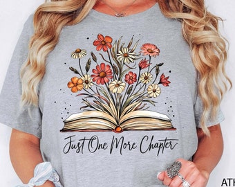 May include: A heather grey t-shirt with a floral design and an open book. The text "Just One More Chapter" is printed on the shirt.