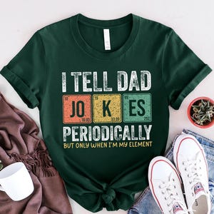 May include: Dark green t-shirt with the text "I TELL DAD JOKES PERIODICALLY BUT ONLY WHEN I'M MY ELEMENT" in white. The word "JOKES" is in a periodic table style. The shirt is paired with white sneakers and blue jeans.
