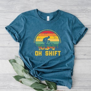 May include: A teal blue t-shirt with a vintage-style graphic of a person riding a bicycle in front of a sunset. The text "OH SHIFT" is printed below the graphic.