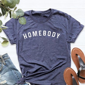 Puede incluir: Una camiseta azul marino con el texto "HOMEBODY" en letras blancas. La camiseta est&aacute; colocada sobre una superficie blanca con un par de sandalias marrones y un par de pantalones vaqueros azules.