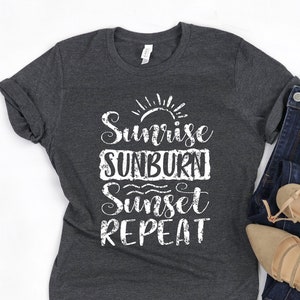 May include: A dark grey t-shirt with white text that reads "Sunrise Sunburn Sunset Repeat". The shirt is laid flat on a white surface with a pair of brown sandals and blue jeans.