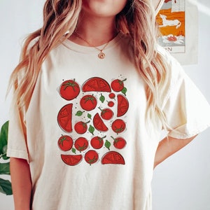 Può includere: Una maglietta bianca oversize con una stampa grafica di pomodori rossi e foglie verdi. La maglietta è indossata da una persona che indossa pantaloncini neri.