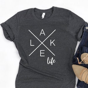 Può includere: Una maglietta grigia scura con un design grafico bianco che dice "LAKE life" con le lettere "LAKE" che formano una croce.