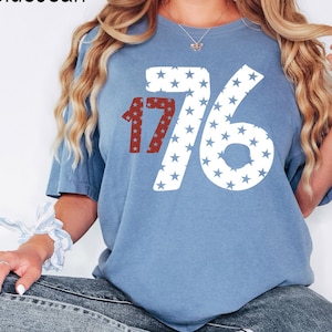Puede incluir: Una camiseta azul con un gr&aacute;fico blanco y rojo del n&uacute;mero 1776. El n&uacute;mero est&aacute; formado por estrellas blancas sobre un fondo azul.
