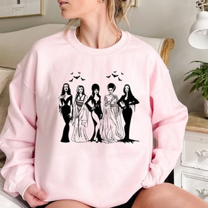 Può includere: Felpa rosa con una grafica in bianco e nero di cinque donne in abiti gotici. Le donne sono in piedi in fila e hanno pipistrelli che volano intorno a loro.