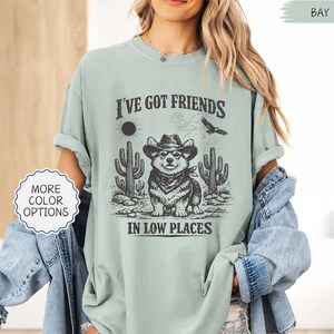 May include: A light green t-shirt with the text "I'VE GOT FRIENDS IN LOW PLACES" and a graphic of a corgi wearing a cowboy hat and bandana, with cacti and a sun in the background. The shirt has rolled-up sleeves.