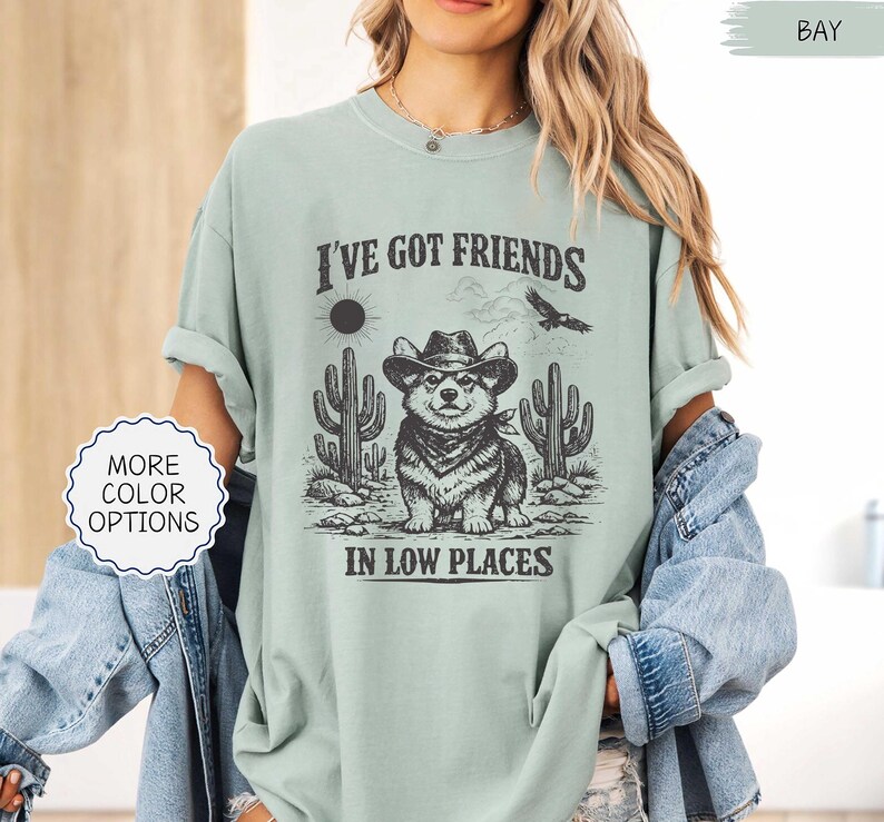 May include: A light green t-shirt with the text "I'VE GOT FRIENDS IN LOW PLACES" and a graphic of a corgi wearing a cowboy hat and bandana, with cacti and a sun in the background. The shirt has rolled-up sleeves.