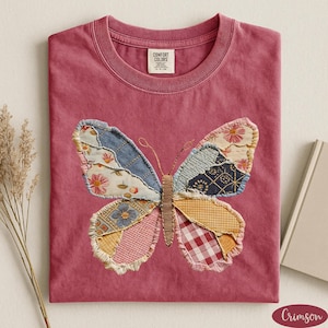 May include: Crimson-colored t-shirt with a crew neck. The shirt features a large butterfly applique made of various fabric patches in floral, gingham, and patterned designs. The butterfly has a stitched outline and detailed antennae.