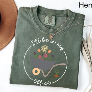 Op de afbeelding: Een groen T-shirt met een witte afbeelding van een kruiwagen vol bloemen en de tekst "Ik ben in mijn kantoor".