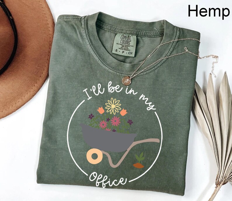 Op de afbeelding: Een groen T-shirt met een witte afbeelding van een kruiwagen vol bloemen en de tekst "Ik ben in mijn kantoor".