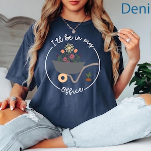 Op de afbeelding: Een marineblauw t-shirt met een witte afbeelding van een kruiwagen vol bloemen. De tekst "I'll be in my office" staat op het shirt gedrukt.