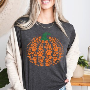 Peut inclure: T-shirt gris fonc&eacute; avec un motif de citrouille en empreintes de pattes orange et une tige verte. La citrouille est compos&eacute;e de nombreuses petites empreintes de pattes. La femme porte le t-shirt et un cardigan cr&egrave;me.